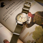 Seagull 1963 flat lay on a printed page, olive NATO strap with polished steel pin buckle and keepers