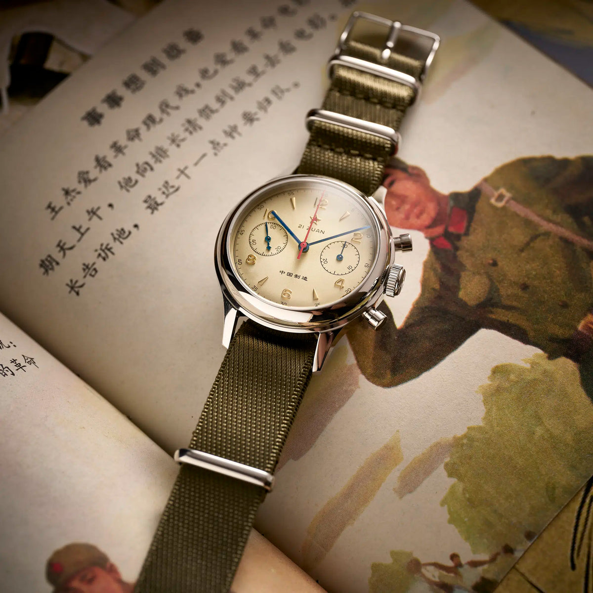 Seagull 1963 flat lay on a printed page, olive NATO strap with polished steel pin buckle and keepers