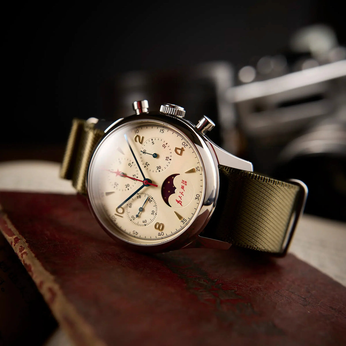 Seagull 1963 Moonphase Cream on its side on a red book, dark background with a vintage camera behind