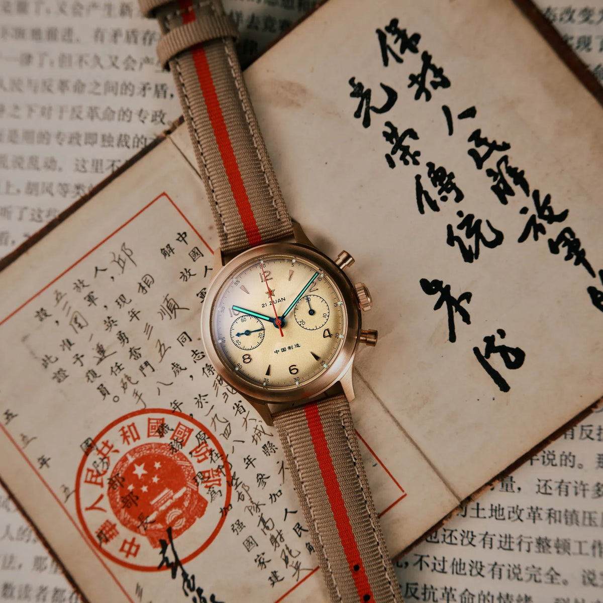 Seagull 1963 Bronze Lume flat lay on Chinese documents with a red seal, grey-beige strap with red stripe